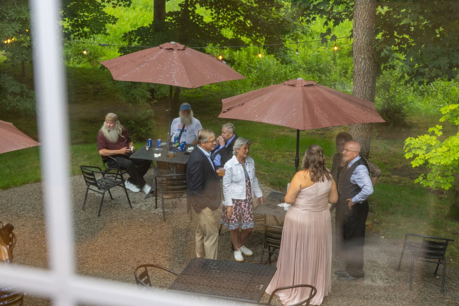 Wedding guests mingle and sit under brown patio umbrellas on an outdoor terrace surrounded by trees.