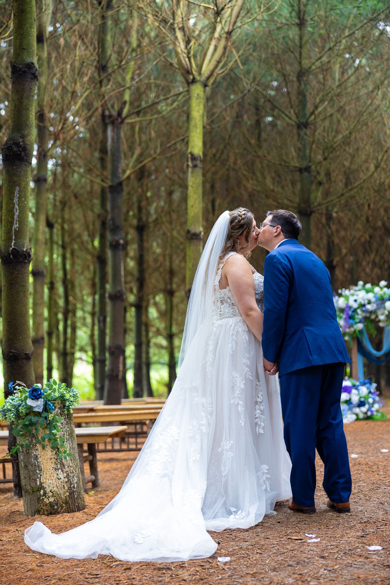 Bride and groom share their first kiss at an outdoor forest wedding ceremony with tall pine trees.