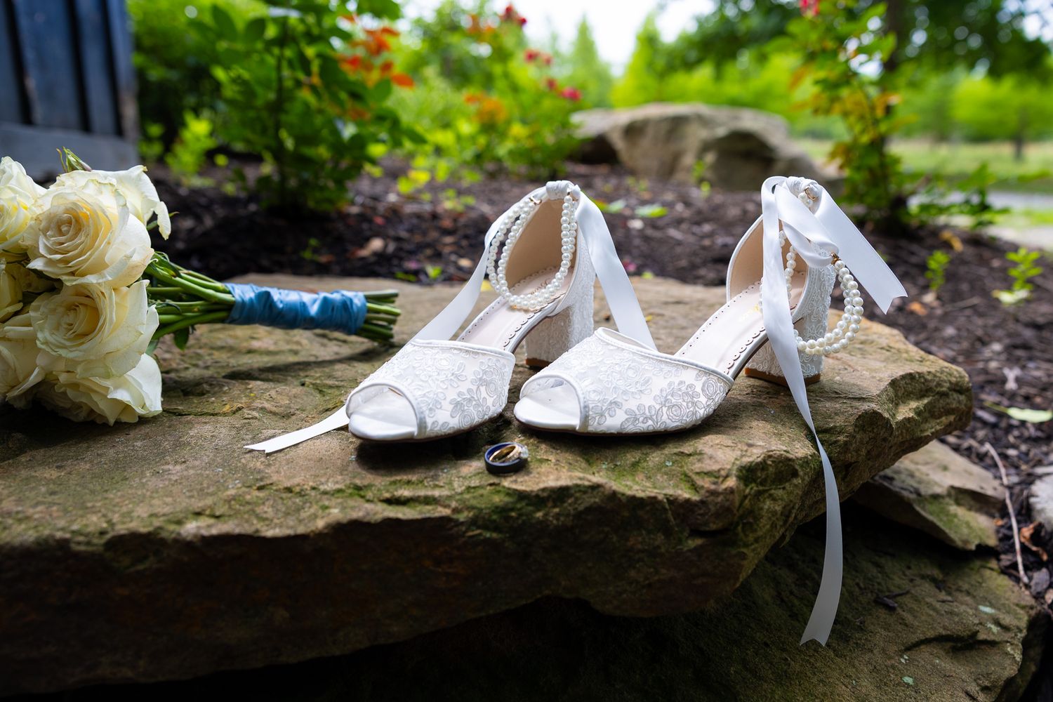 White lace bridal heels with ribbon ties displayed on stone steps beside a bouquet of white roses and a wedding ring.