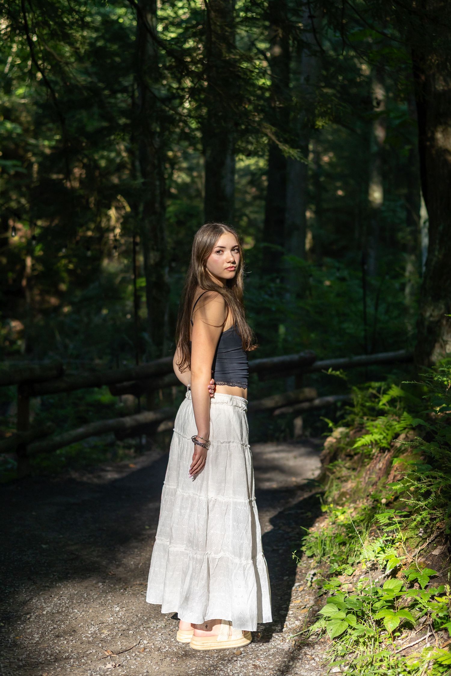 Young woman in white maxi skirt and gray crop top standing on a forest path surrounded by lush green trees.