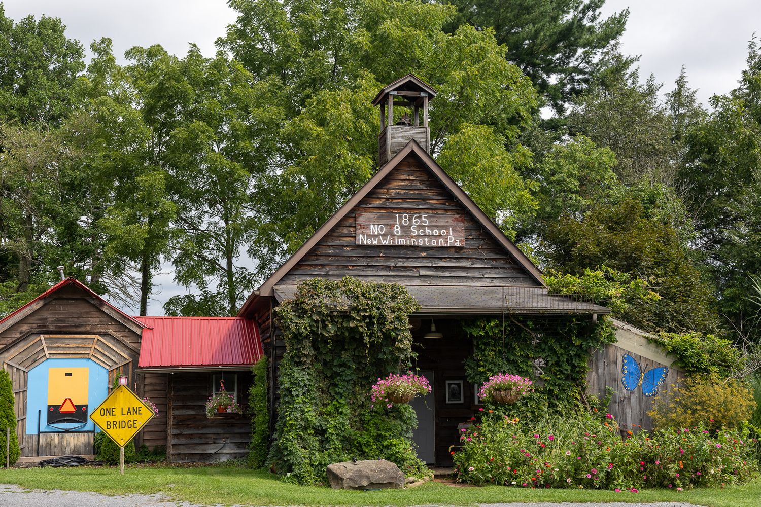Rustic wooden schoolhouse covered in ivy with a bell tower, red roof, and colorful signs amid lush green trees.