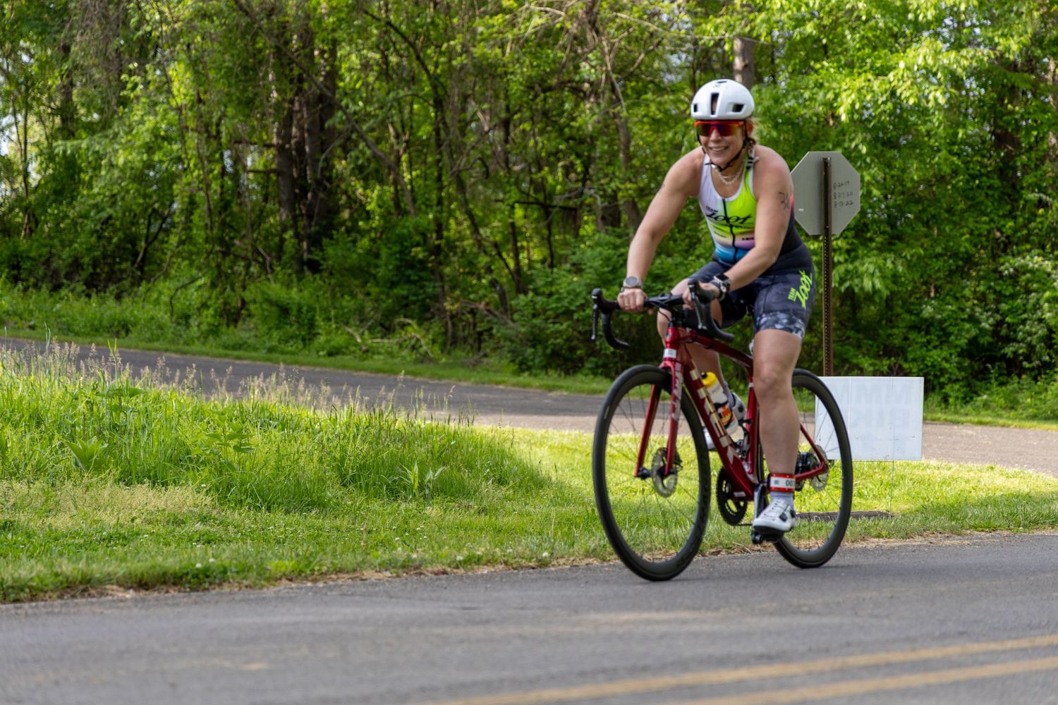 A cyclist in a white helmet and athletic wear rides a red road bike on a paved path surrounded by lush green trees.