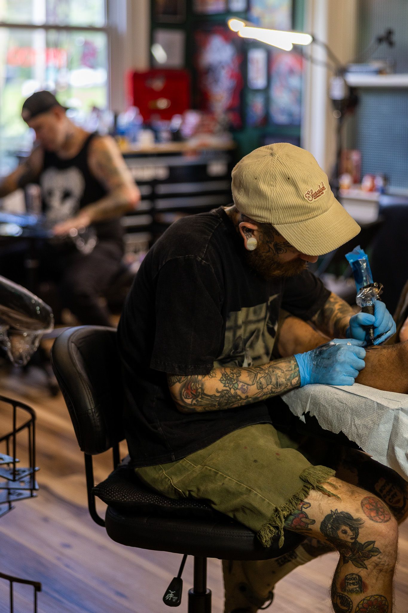 Tattooed artist in cap and gloves carefully tattooing client in busy professional tattoo parlor.