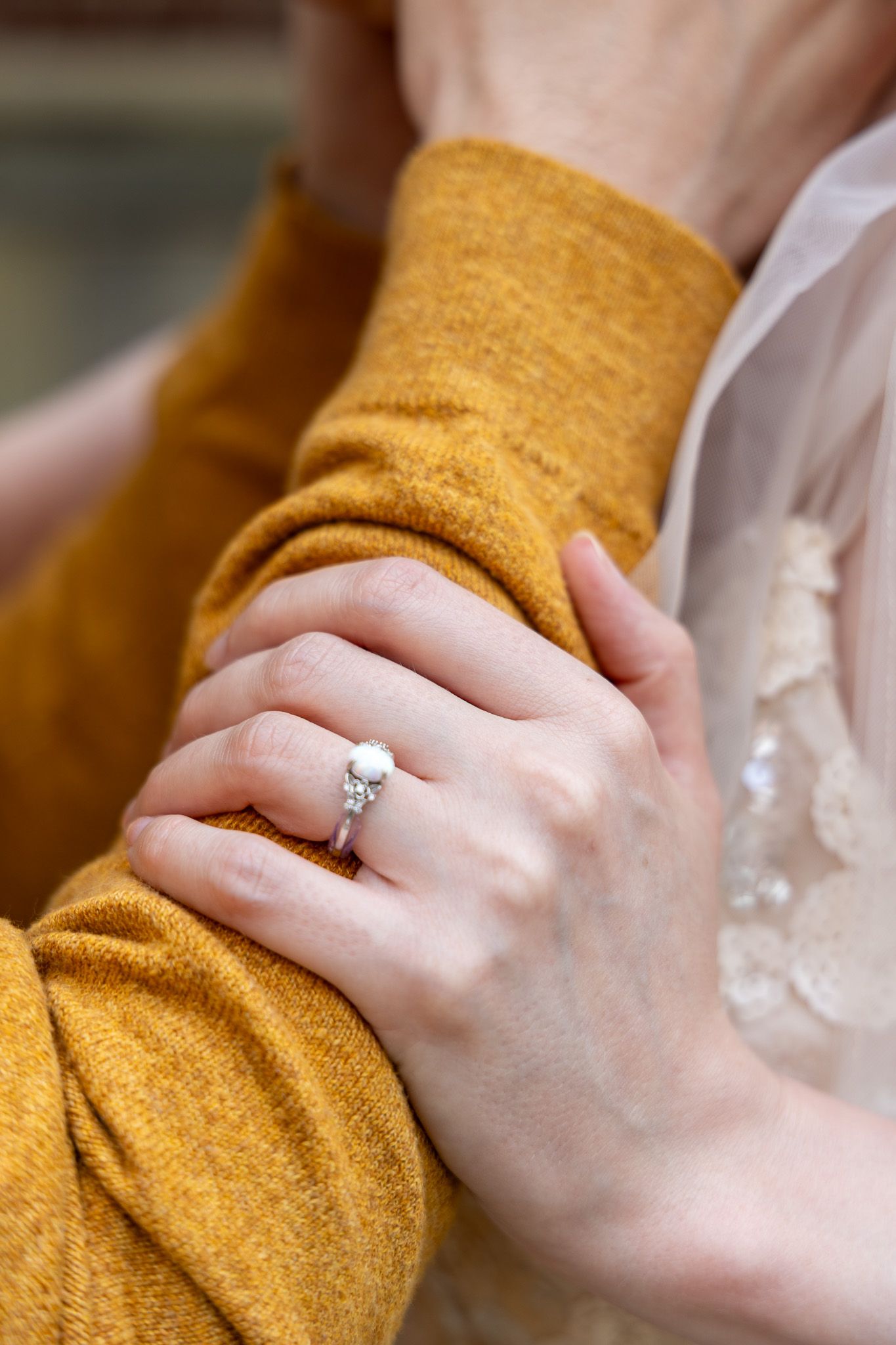 Close-up of a woman's hand wearing a diamond engagement ring, resting on a man's arm in a mustard yellow sweater.
