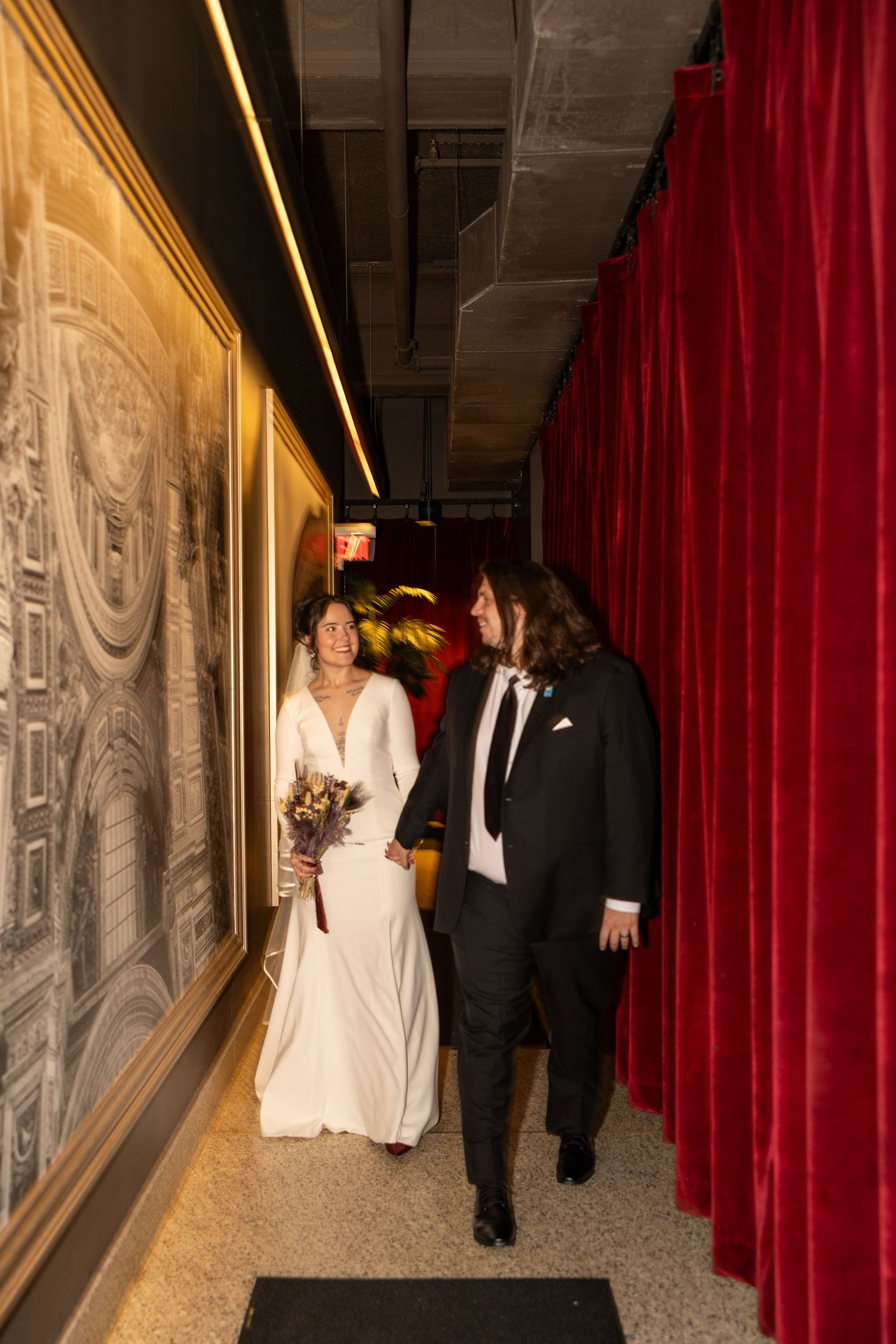 Bride and groom walk hand-in-hand down a narrow hallway lined with red velvet curtains and artwork.