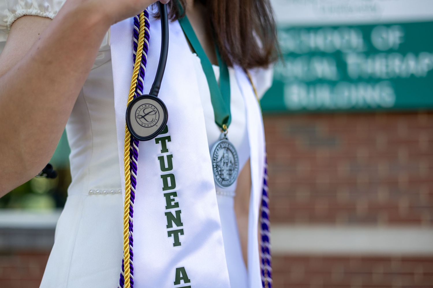 Close-up of graduate wearing Student Athlete stole and medal near Physical Therapy Building sign.