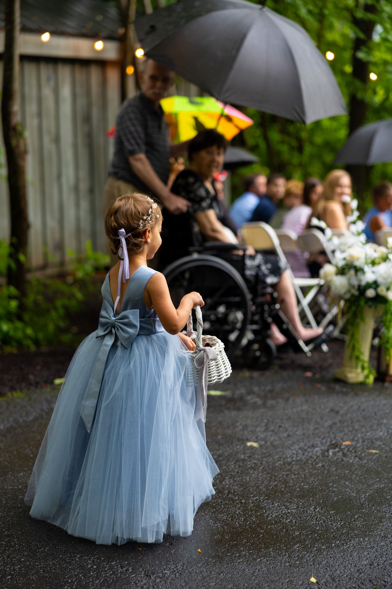 Flower girl in blue tulle dress walks down rainy outdoor wedding aisle carrying white basket.