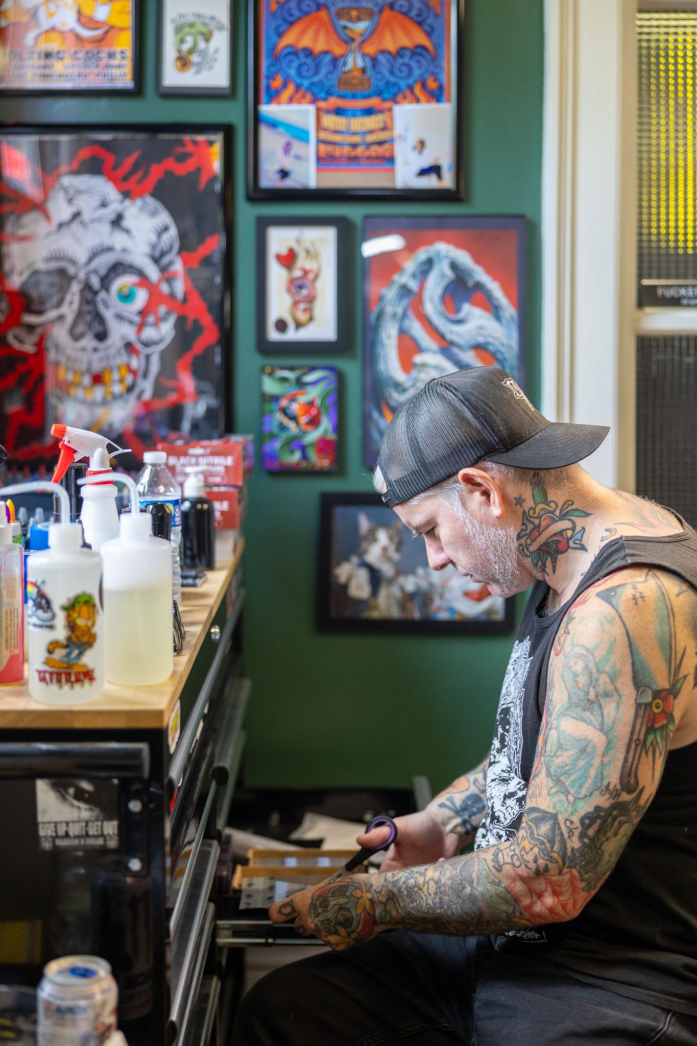 Heavily tattooed artist in a black cap prepares in a vibrant tattoo studio surrounded by colorful skull and art prints.