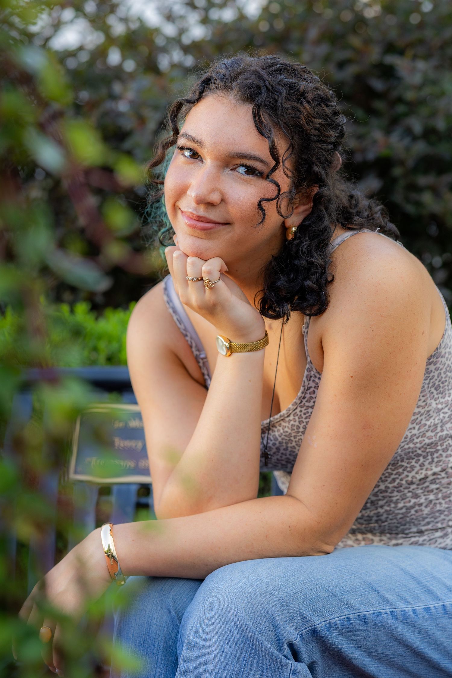 Young woman with curly hair sits outdoors, smiling thoughtfully, wearing a leopard print top, jeans, and gold jewelry.