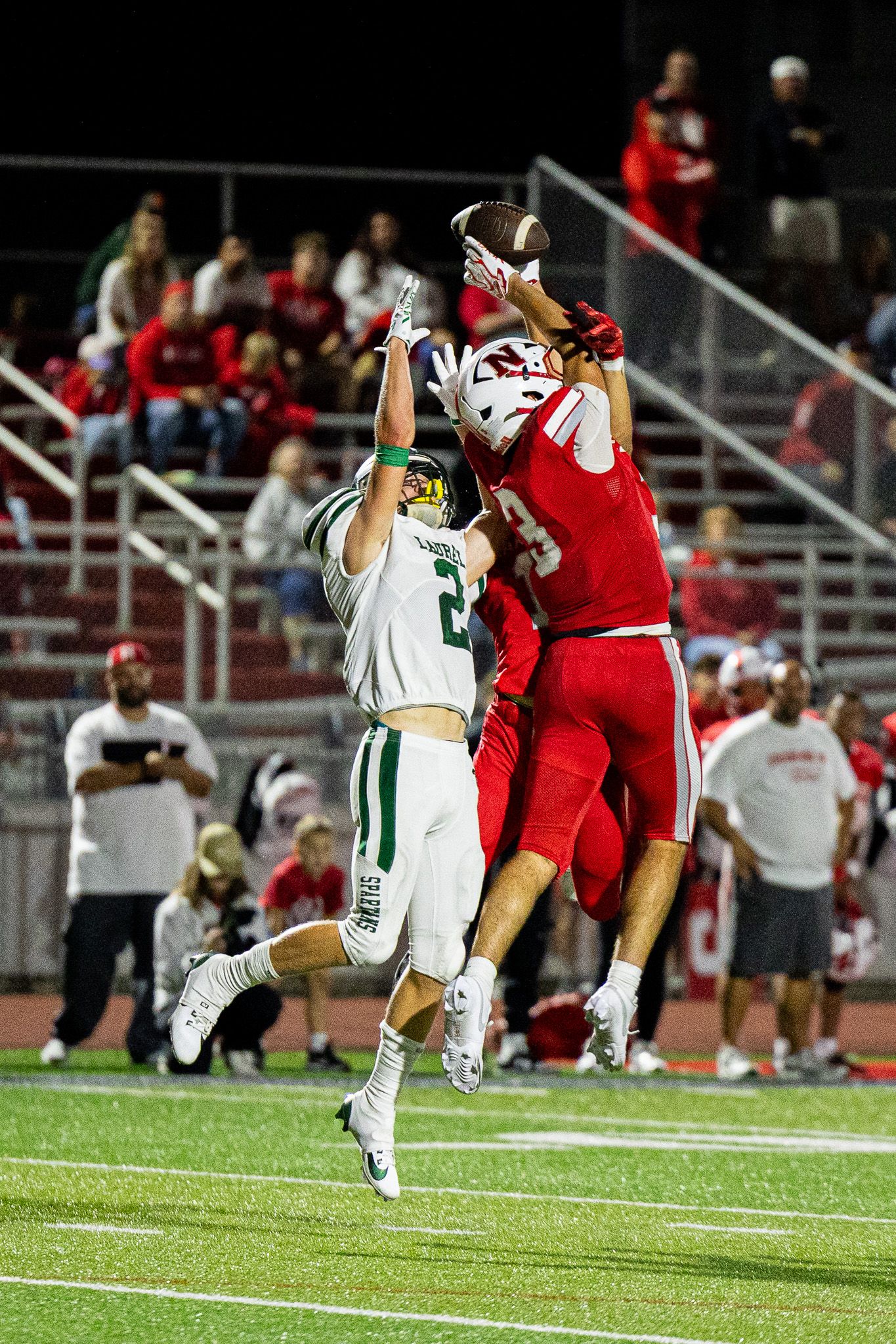 Neshannock football player leaps to catch ball over green Laurel defender during night game at stadium.