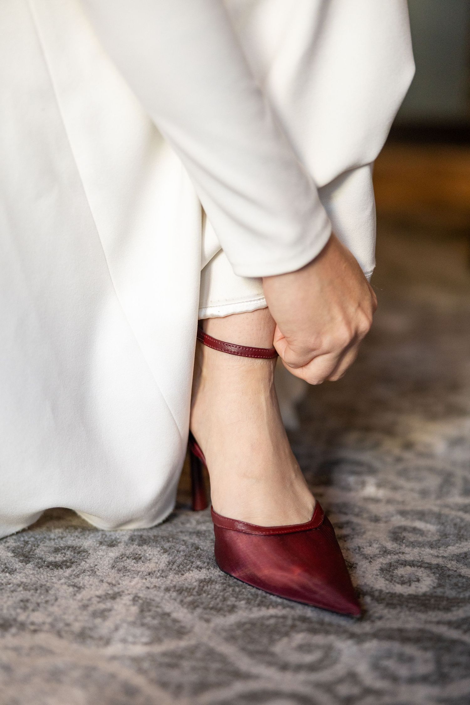 Bride in white dress fastening a deep red pointed-toe heel with ankle strap on patterned carpet.