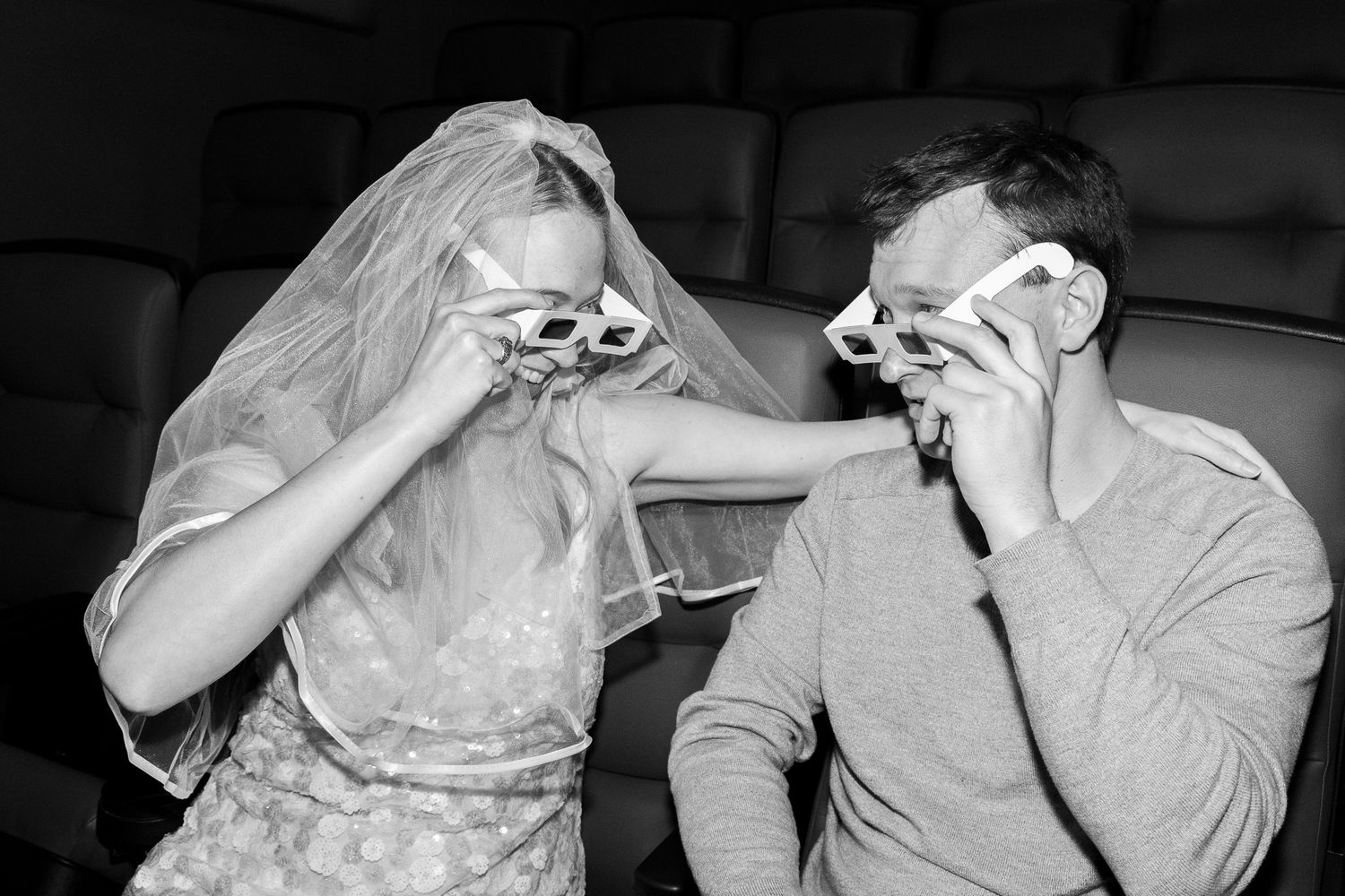 Bride and groom wearing 3D glasses at a movie theater on their wedding day, black and white photo.