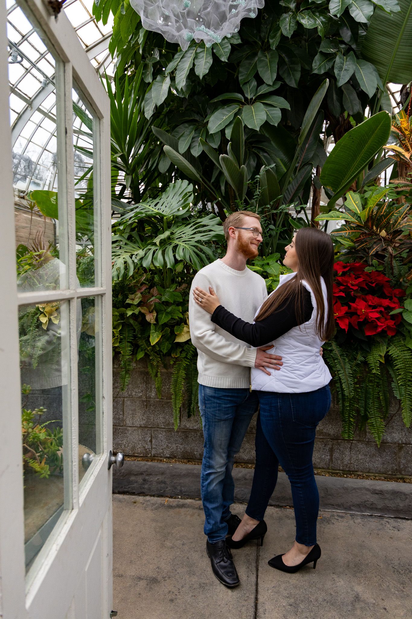 Couple embracing and smiling at each other inside a lush greenhouse surrounded by tropical plants and poinsettias.