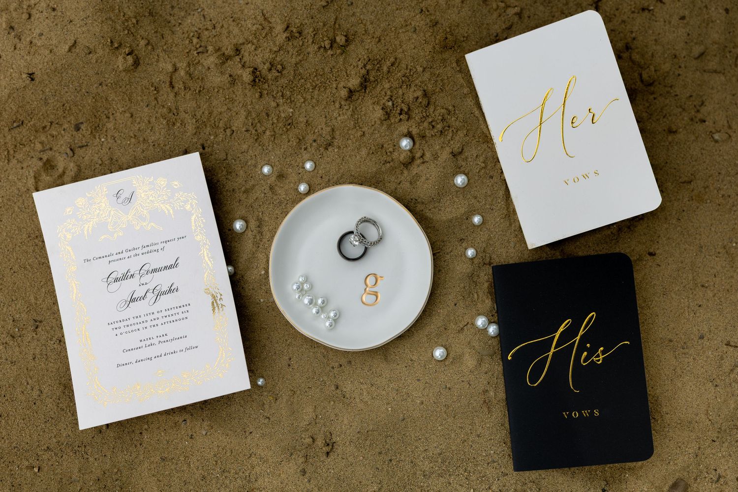 Wedding flat lay with invitation, His and Her vow books, ring dish with bands, and pearl accents on sandy surface.