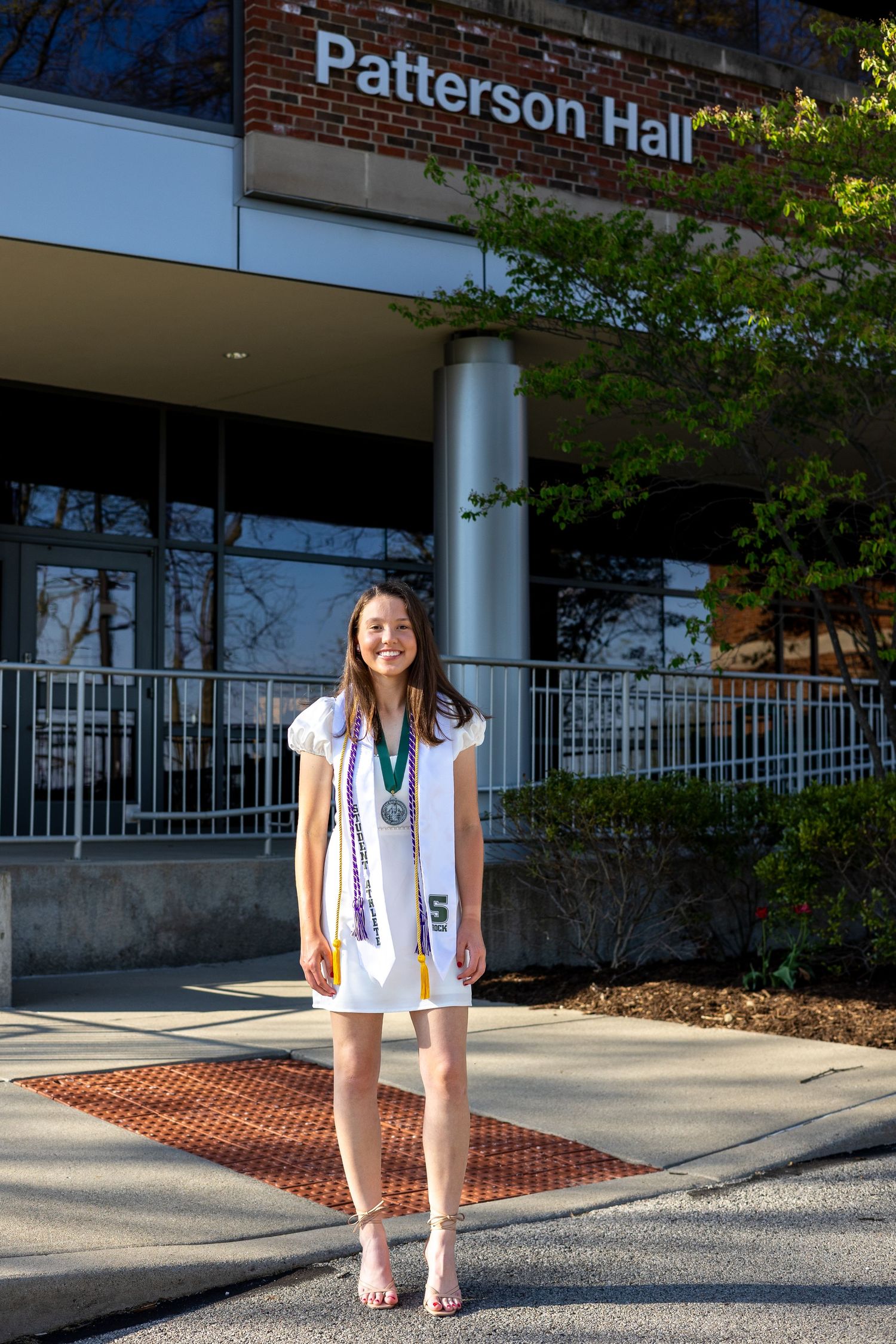 Graduate in white dress with medal stands before Patterson Hall at Slippery Rock University.