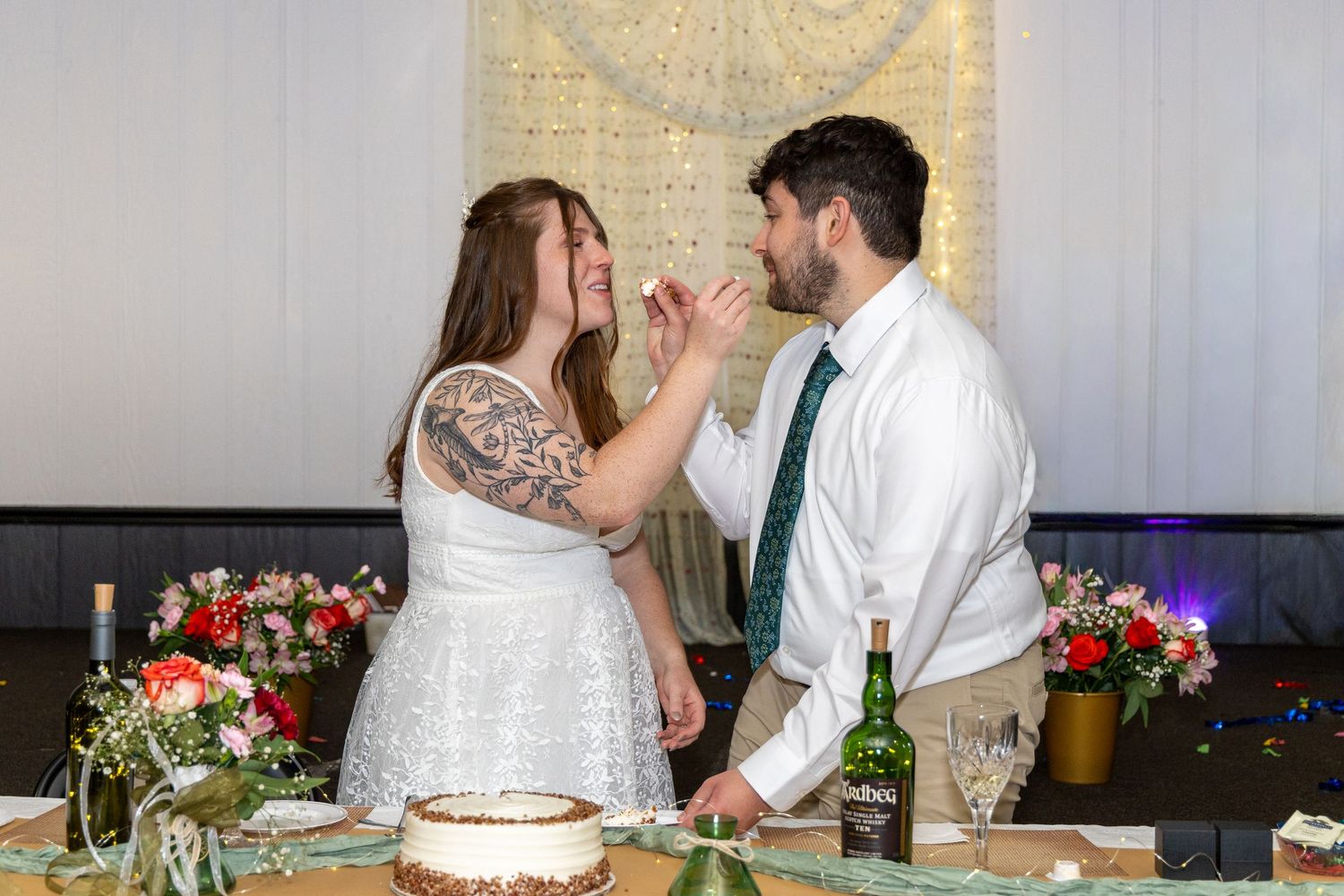 Bride and groom share wedding cake at reception table decorated with colorful flowers and greenery.