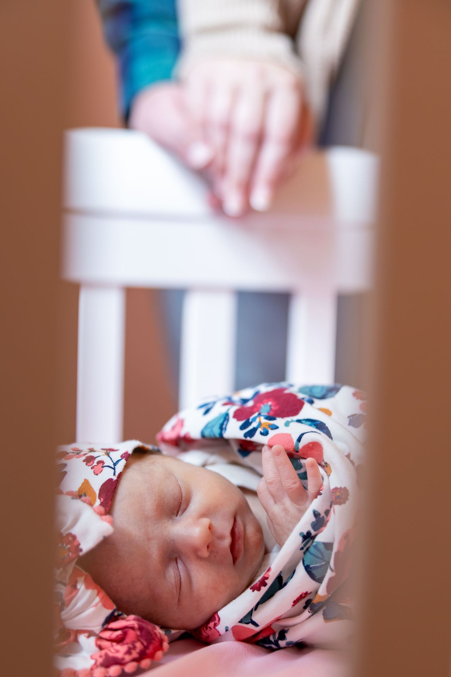 Newborn baby sleeping peacefully in a white crib, wrapped in floral blanket, with parent's hands resting on the rail.