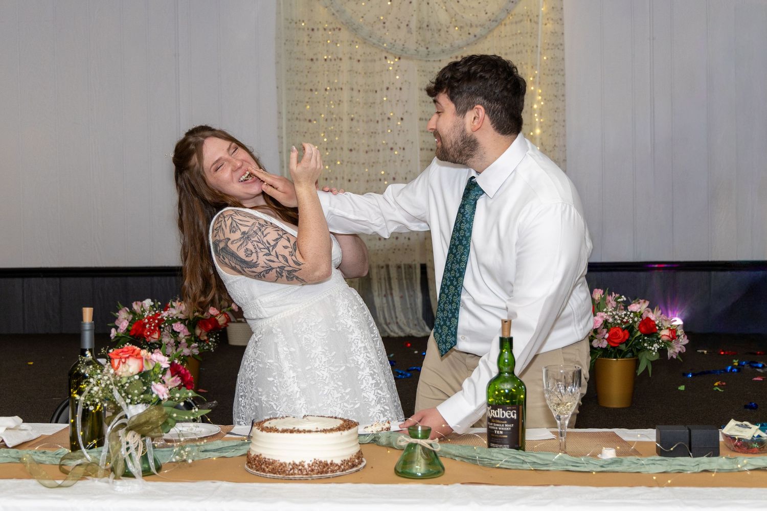 Bride and groom smiling as groom playfully feeds wedding cake to bride at decorated reception table.