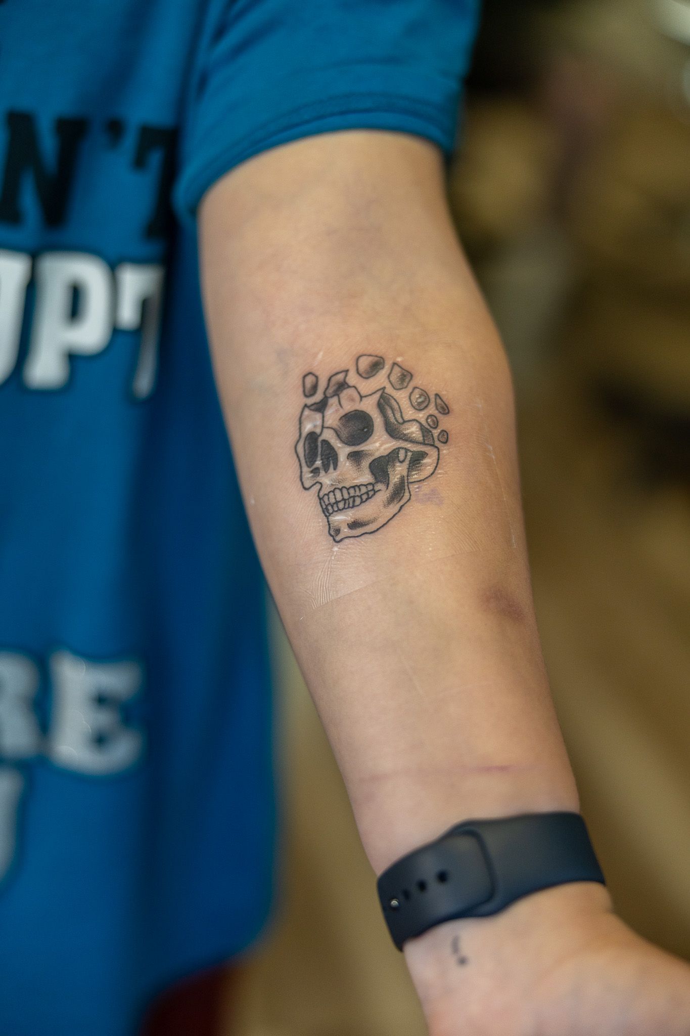 Small black and grey skull tattoo on a person's forearm wearing a blue shirt and fitness tracker.