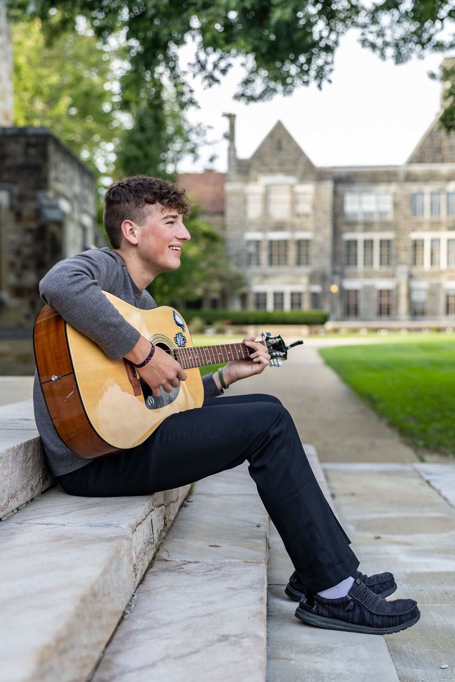 Young man smiling while playing acoustic guitar on stone steps outside a historic building.