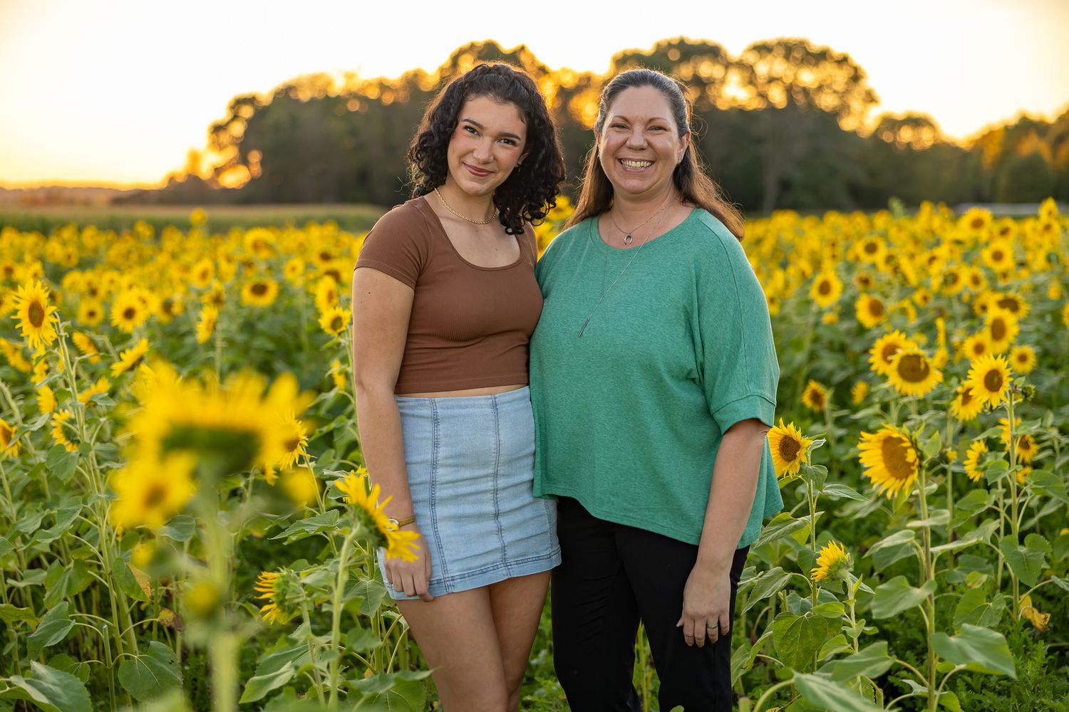 Two women smiling in a sunflower field at golden hour sunset.