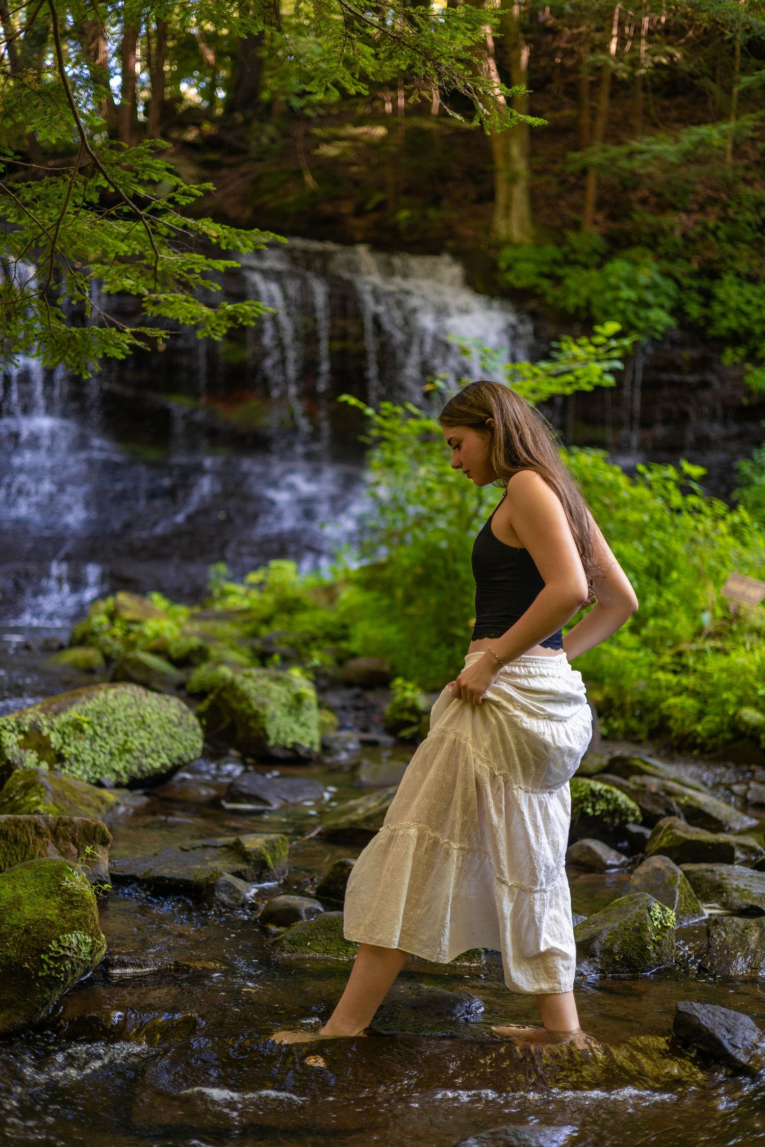 Young woman in white skirt wading barefoot in a rocky stream with a lush green waterfall backdrop.