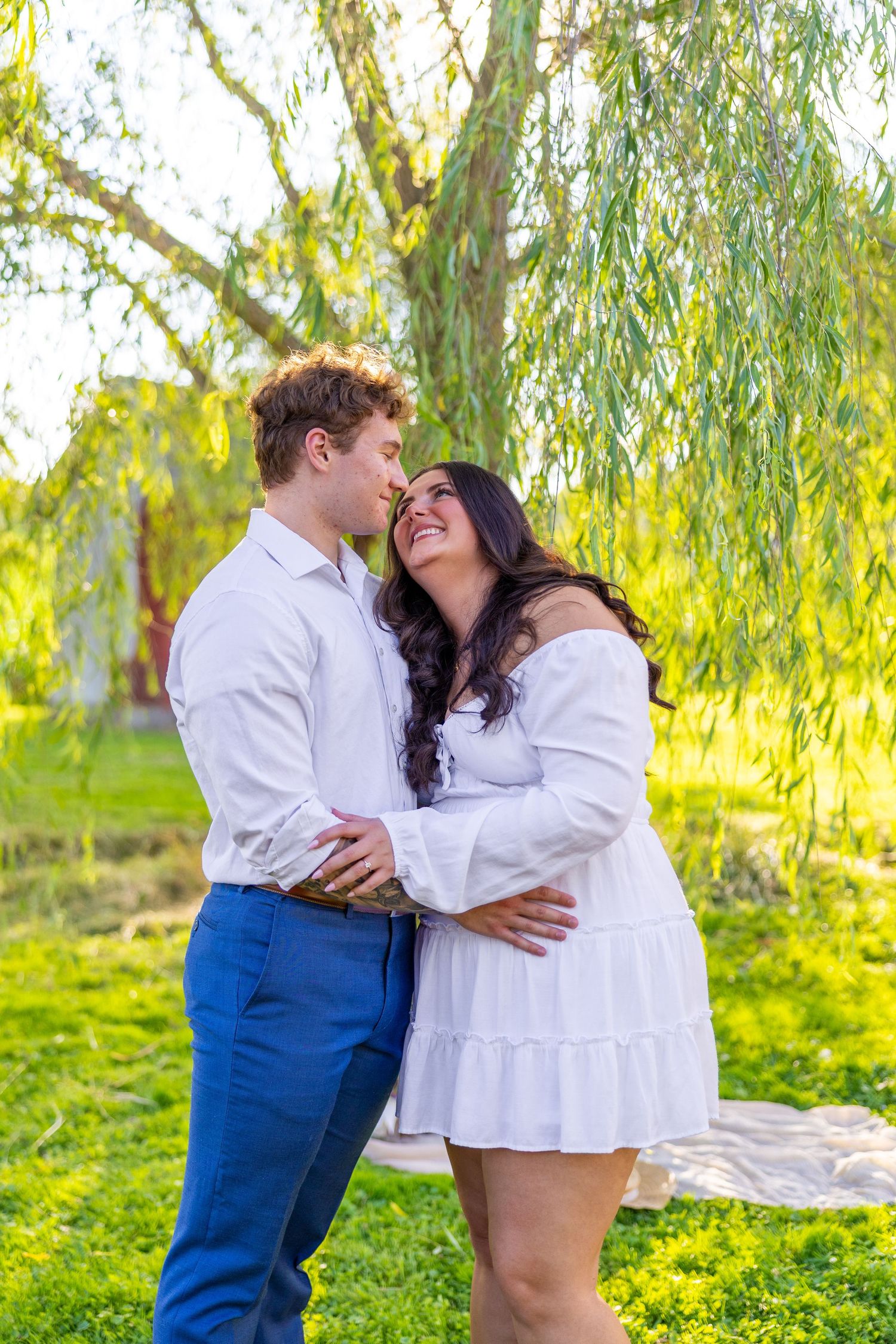 Couple shares a tender almost-kiss embrace under sunlit weeping willow tree during engagement session.