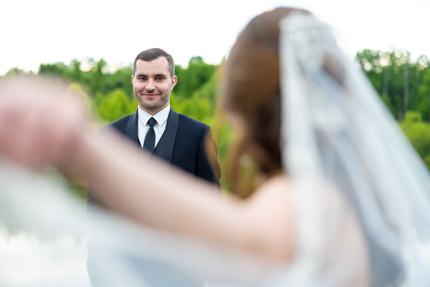 Groom gazes lovingly at bride with veil blowing in foreground during outdoor wedding portrait.