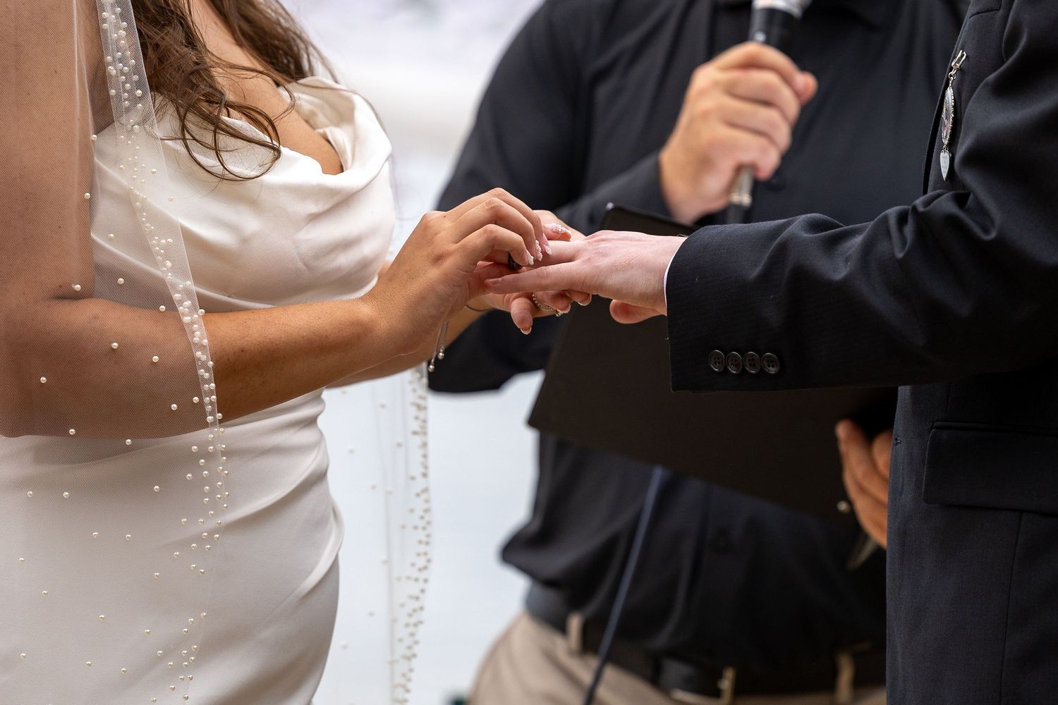 Groom placing wedding ring on bride's finger during ceremony, officiant holding microphone in background.