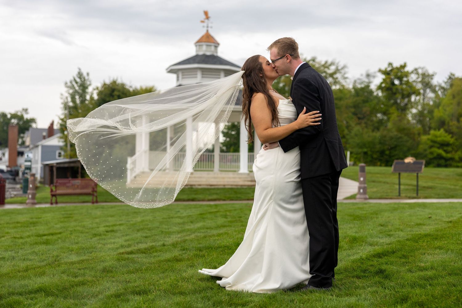 Bride and groom share a romantic kiss outdoors, with veil blowing in wind, lush green lawn and gazebo in background.