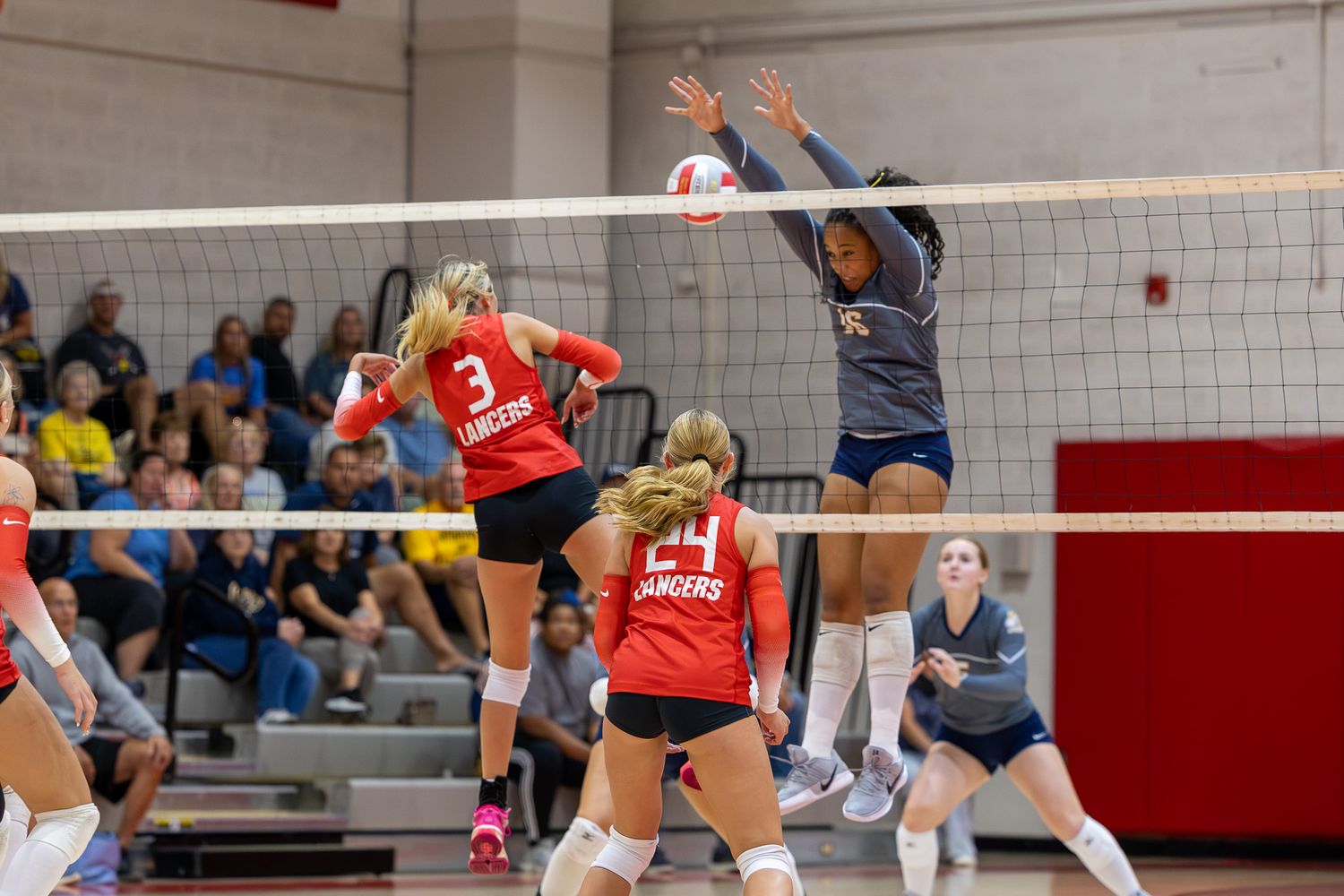 Female volleyball players compete at the net, with a player in red jersey #3 spiking against a defender in navy blue.