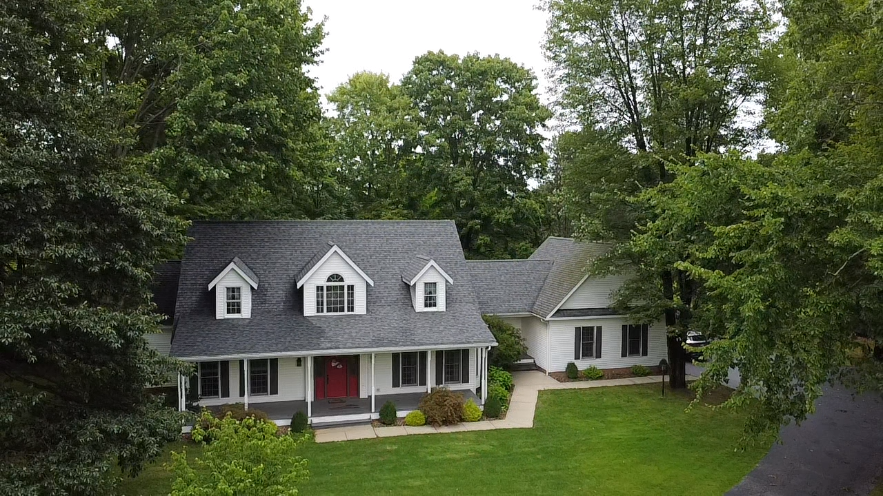 Aerial view of a charming Cape Cod home with gray roof, red door, white trim, and lush surrounding trees.