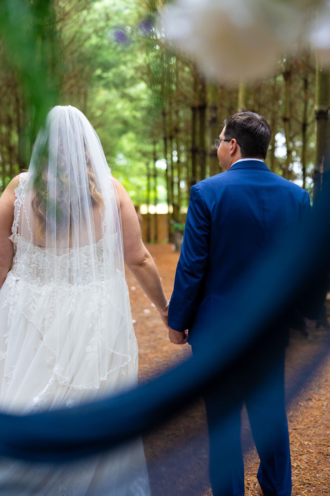 Bride and groom walk hand in hand through a lush forest path, viewed from behind, showcasing her white gown and veil.