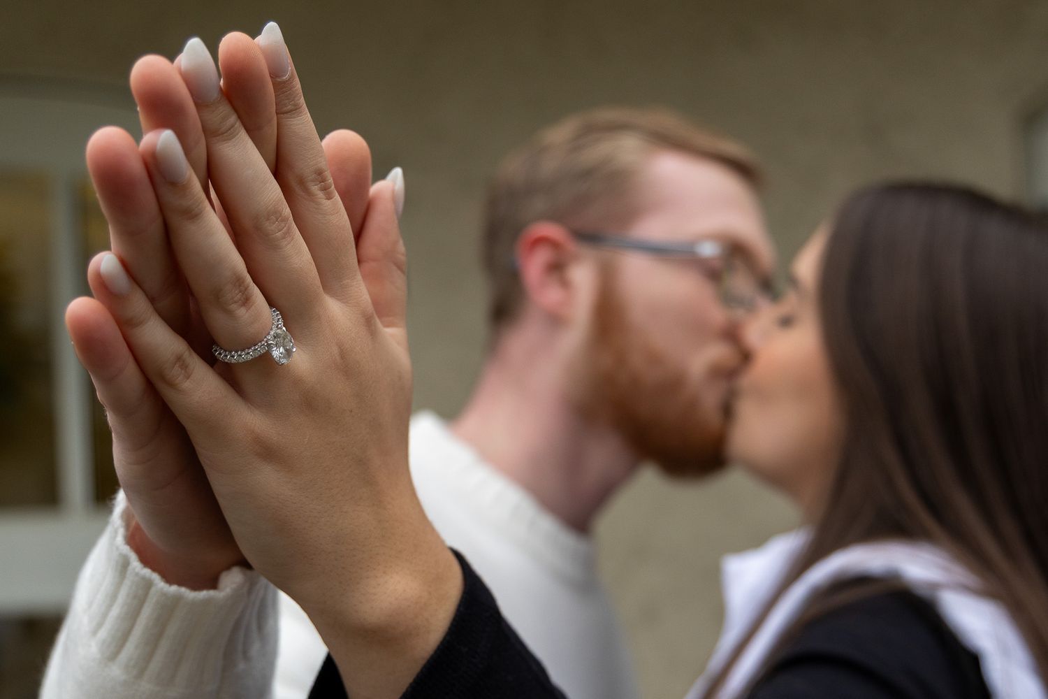 Couple kissing in background while showing off diamond engagement ring on hand in foreground.