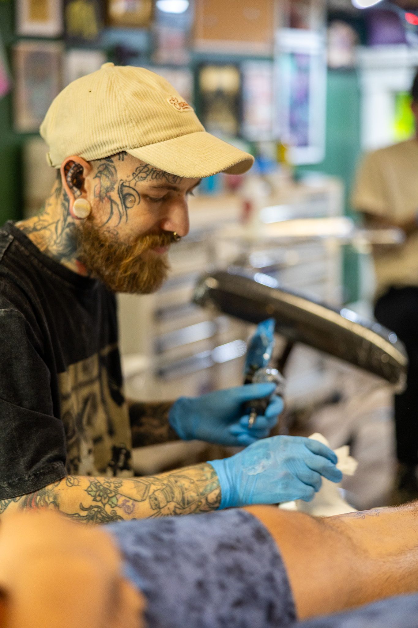 Bearded tattoo artist with neck tattoos and cap carefully tattoos a client's arm wearing blue gloves.