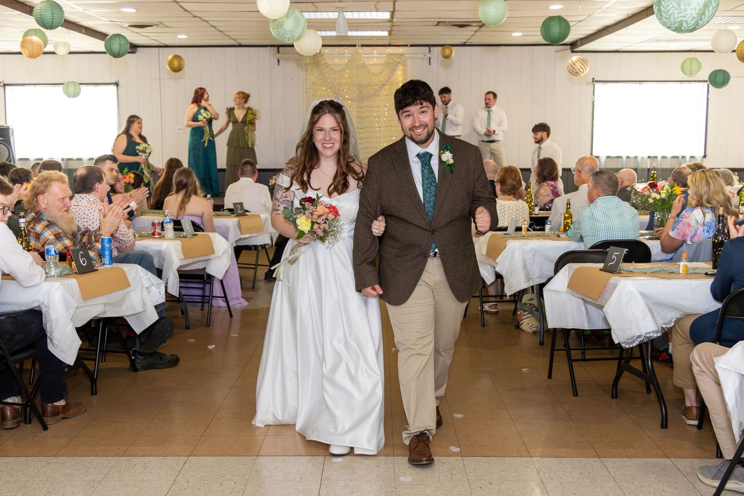 Bride and groom walk hand-in-hand through wedding reception hall with guests applauding and decorated tables in background.