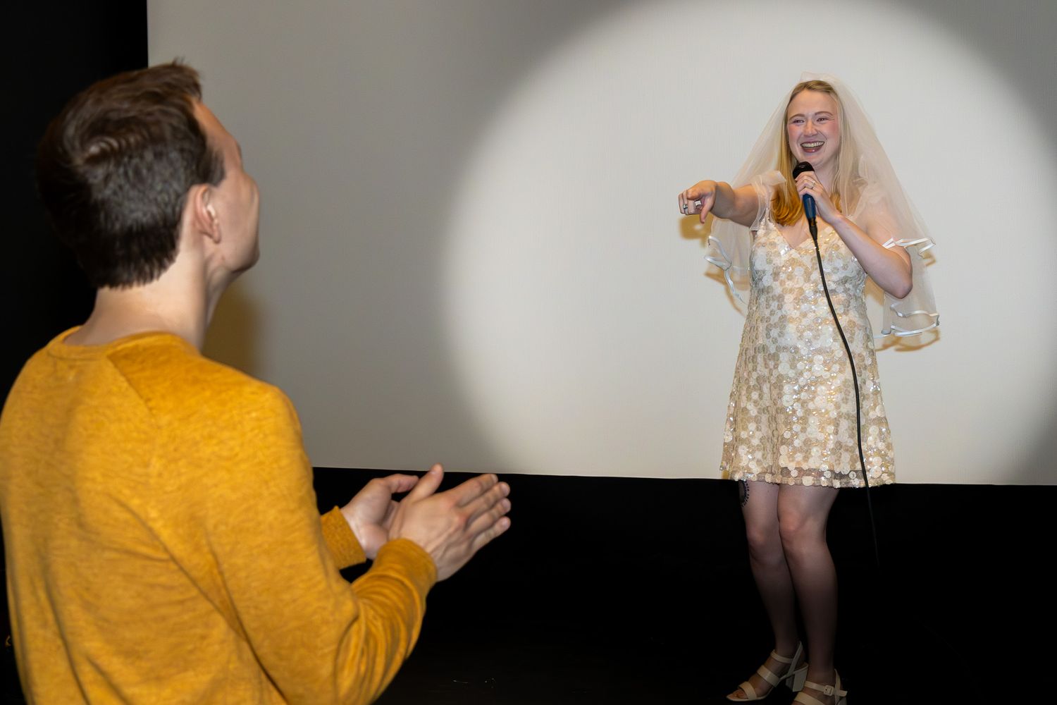 Blonde woman in floral dress speaking at microphone while man in mustard sweater applauds.