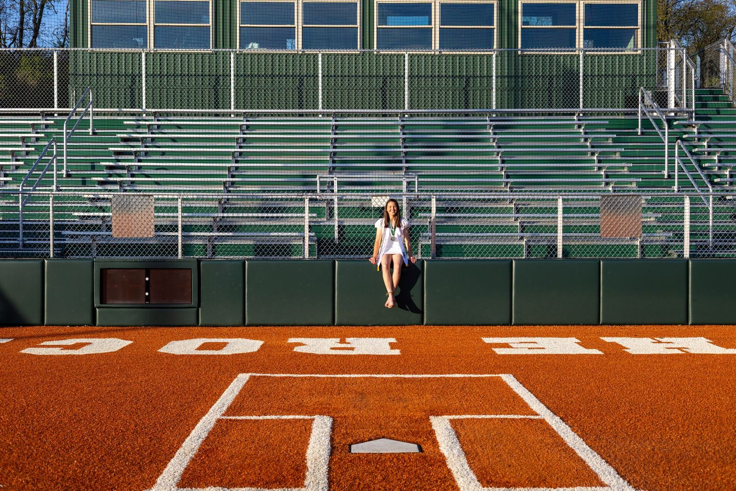Young woman in white outfit standing alone on red turf baseball field in front of green bleachers.
