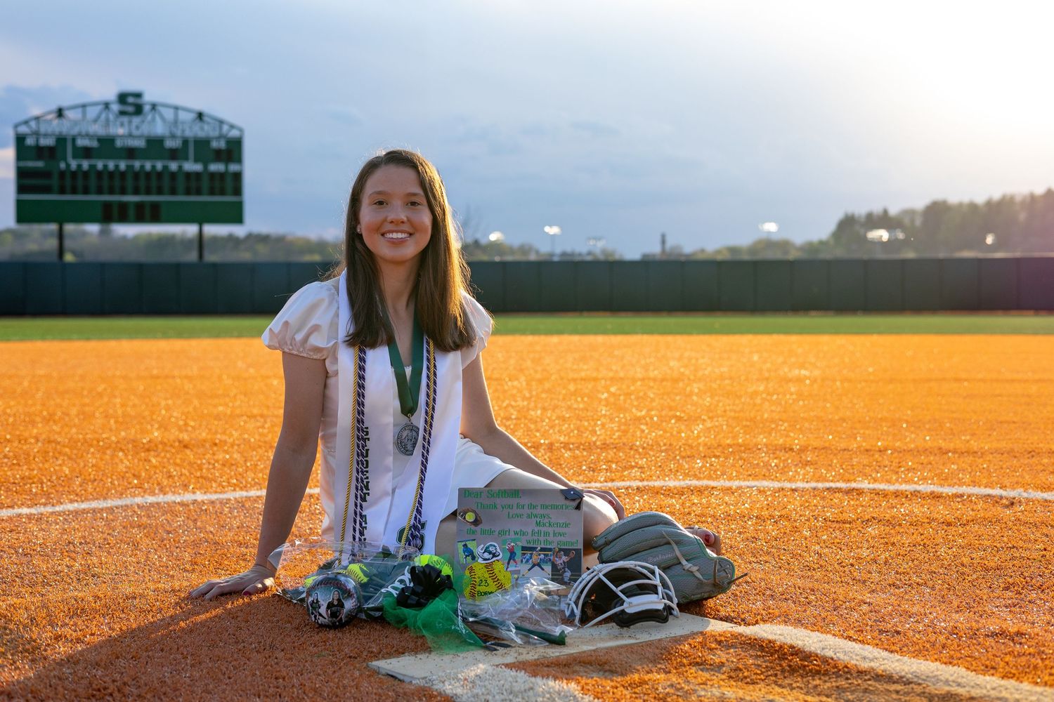 Smiling female graduate sits on baseball field holding softball with glove and awards displayed nearby.