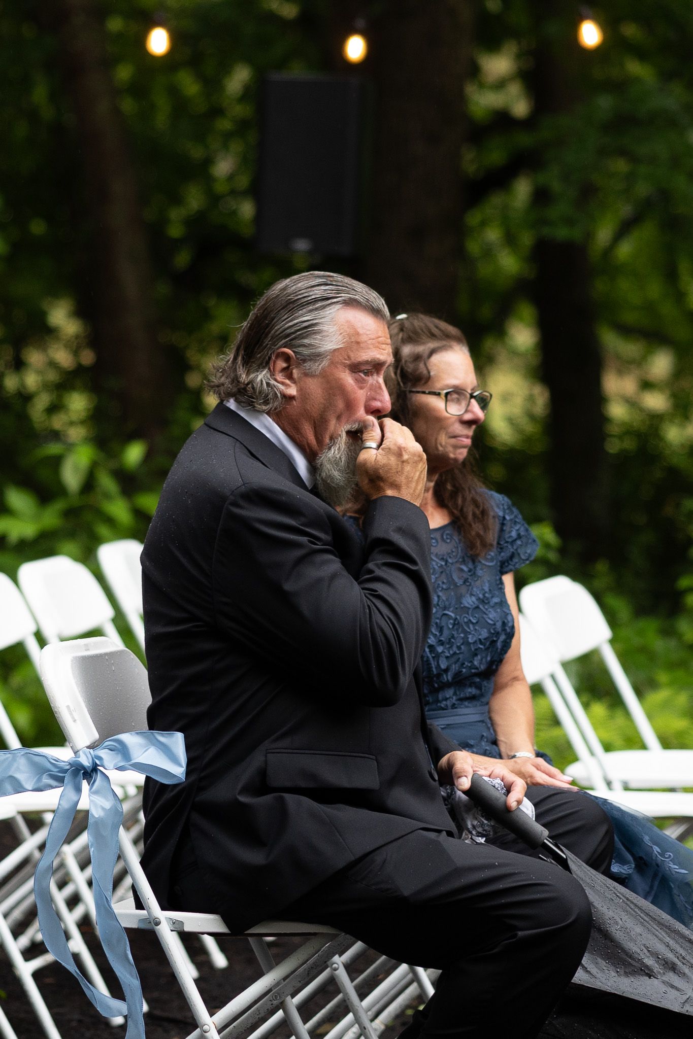 Emotional wedding guest in black suit wipes tears while watching outdoor ceremony from white folding chair.