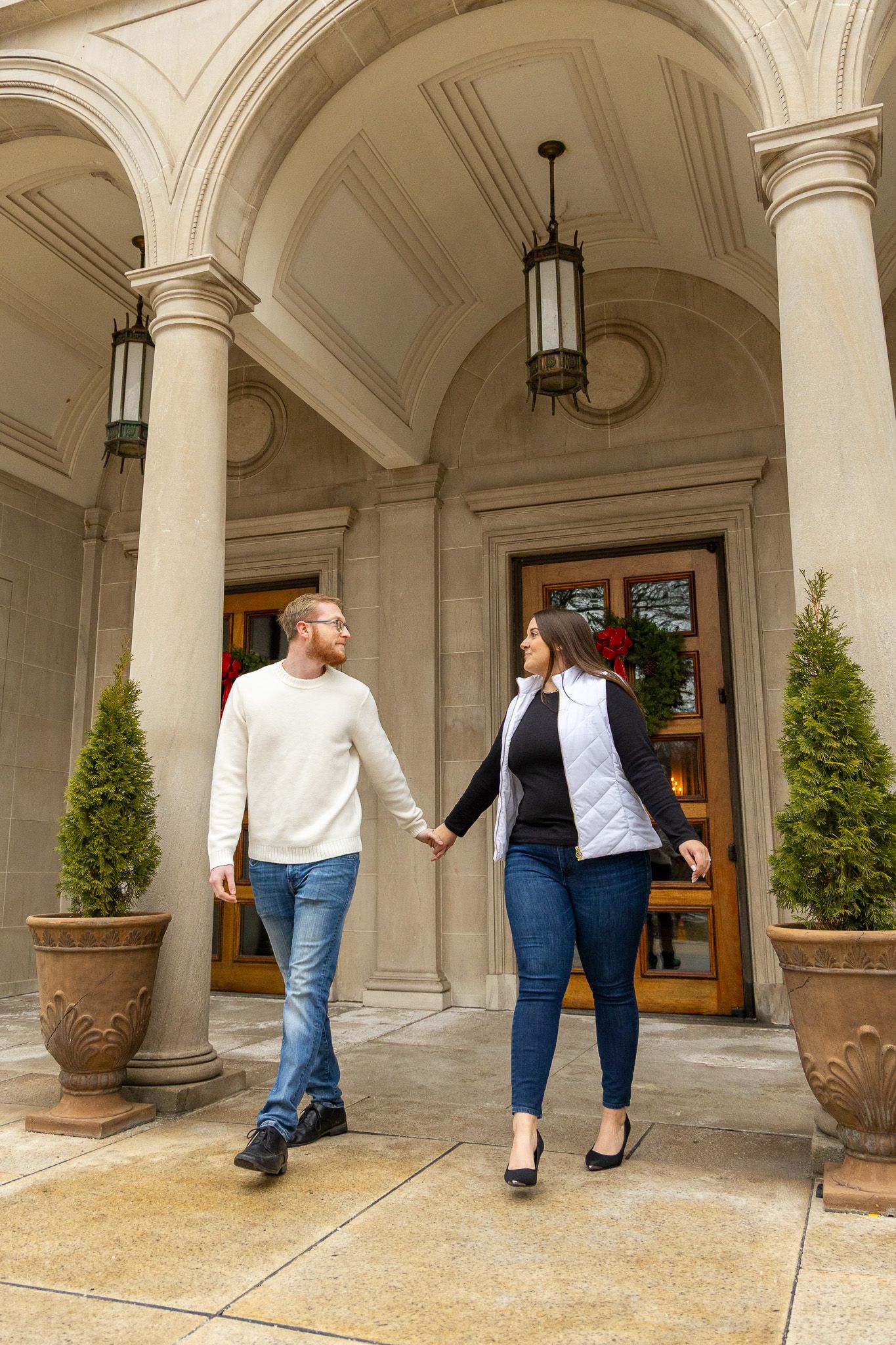 Couple holding hands walking under elegant stone archway with columns and holiday wreath decorations.