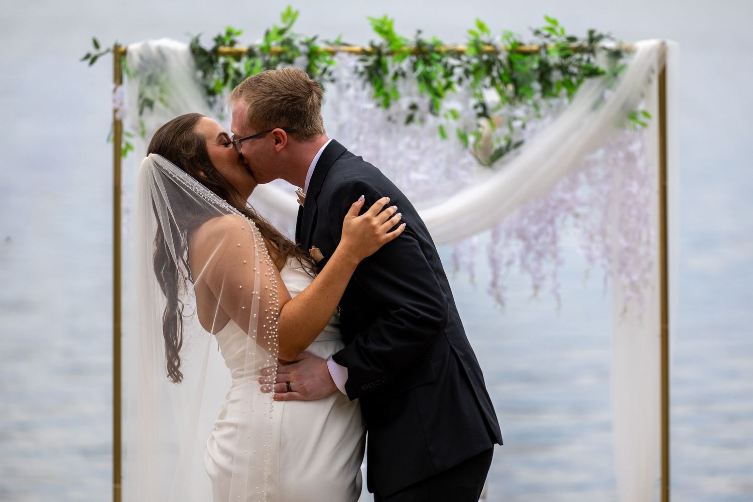 Bride and groom share first kiss at outdoor waterfront wedding ceremony under floral arch with greenery.