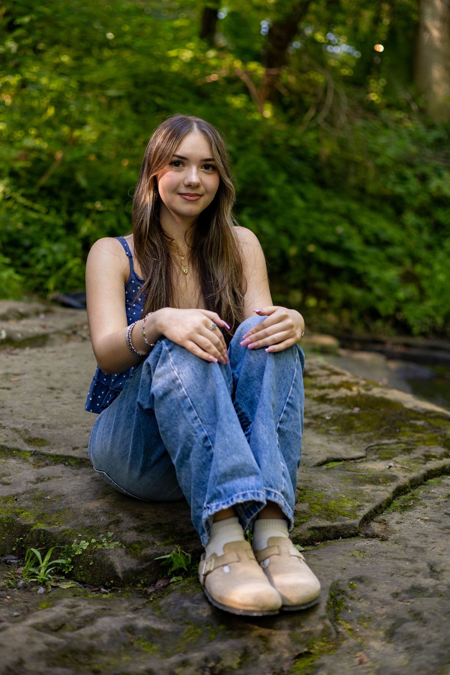 Young woman in blue tank top and jeans sitting on stone steps surrounded by lush green trees.