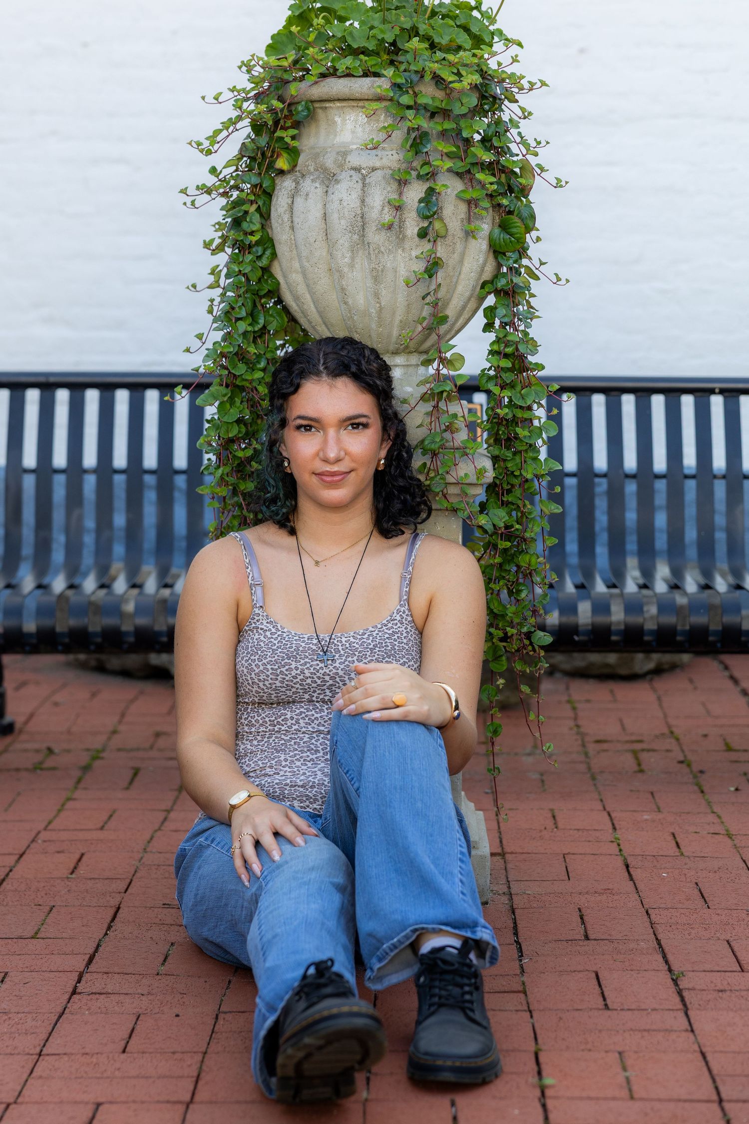 Young woman sitting on brick ground in jeans and floral top, posing in front of a decorative urn with ivy.