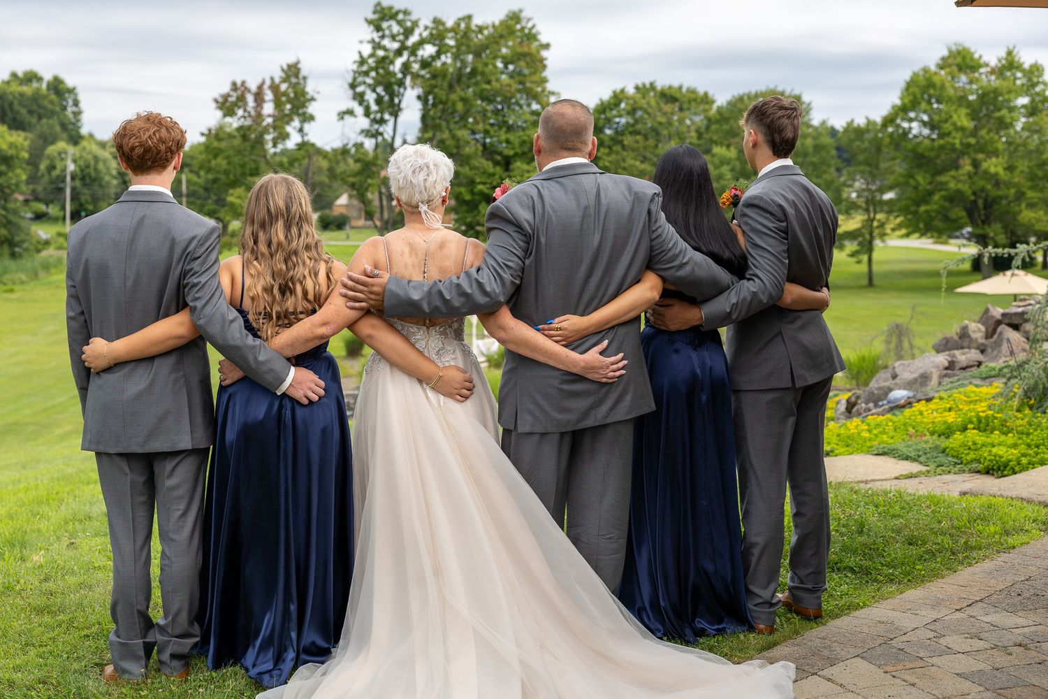 Wedding party poses with backs to camera, bridesmaids in navy blue gowns flanking bride and groom outdoors.