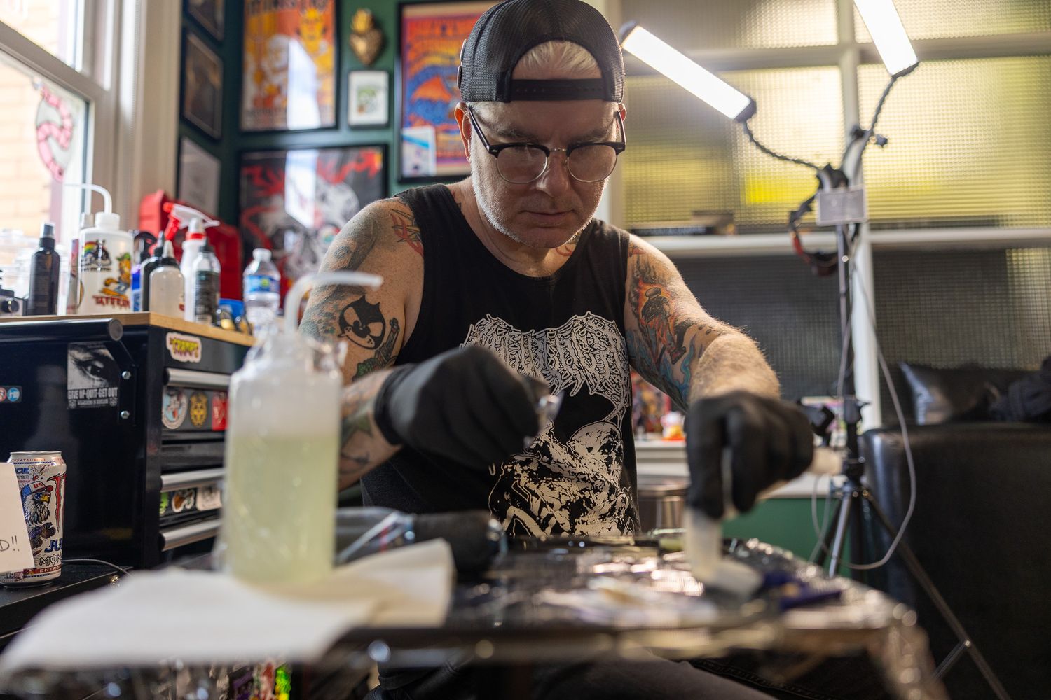 Heavily tattooed artist in black gloves preparing tattoo equipment at a cluttered studio workstation.