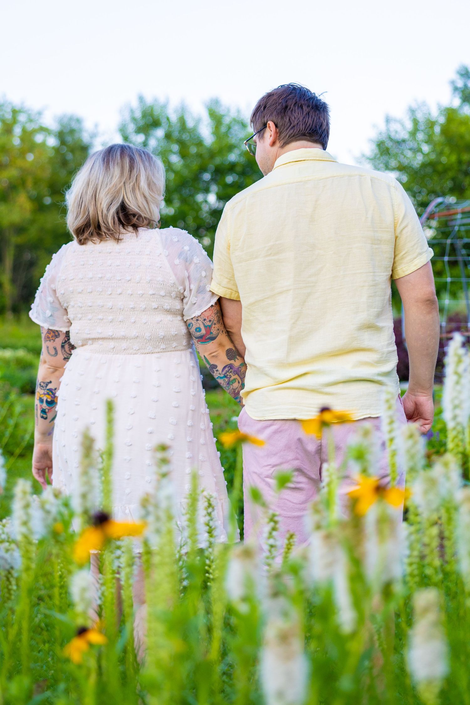 A couple with tattoos walks hand in hand through a colorful wildflower garden, viewed from behind.