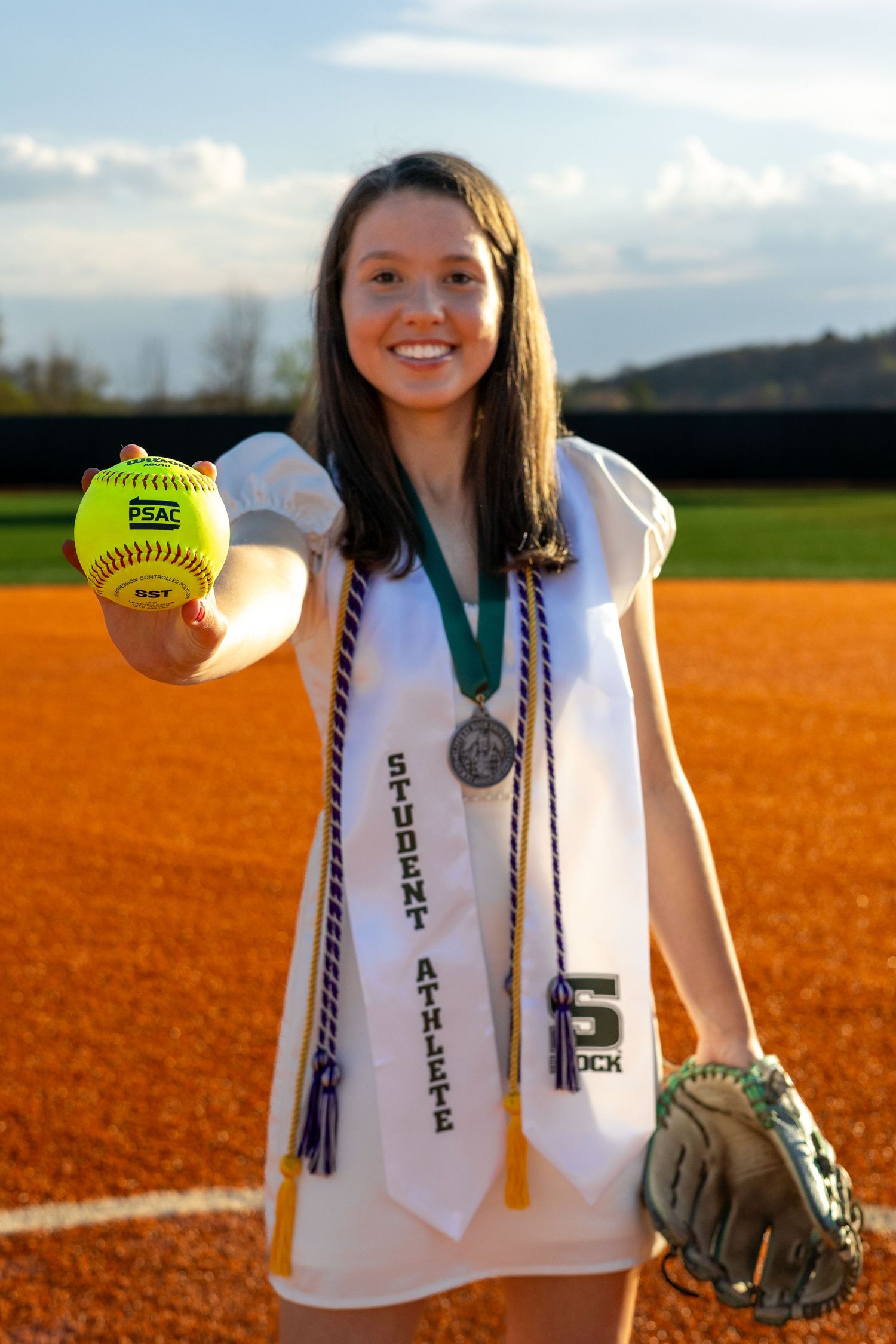 Female student athlete holds softball toward camera wearing graduation medal and Student Athlete sash.