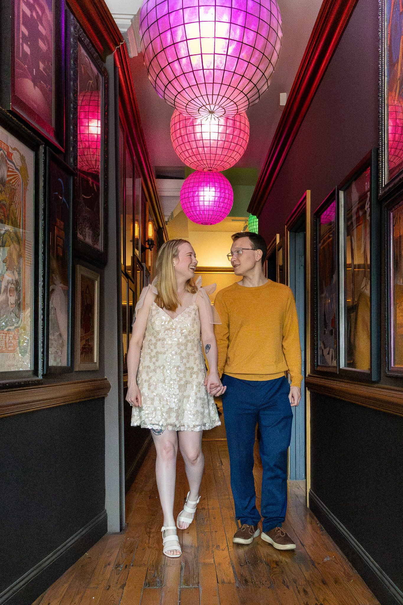 Couple holding hands walking through colorful hallway with pink disco ball lights overhead.