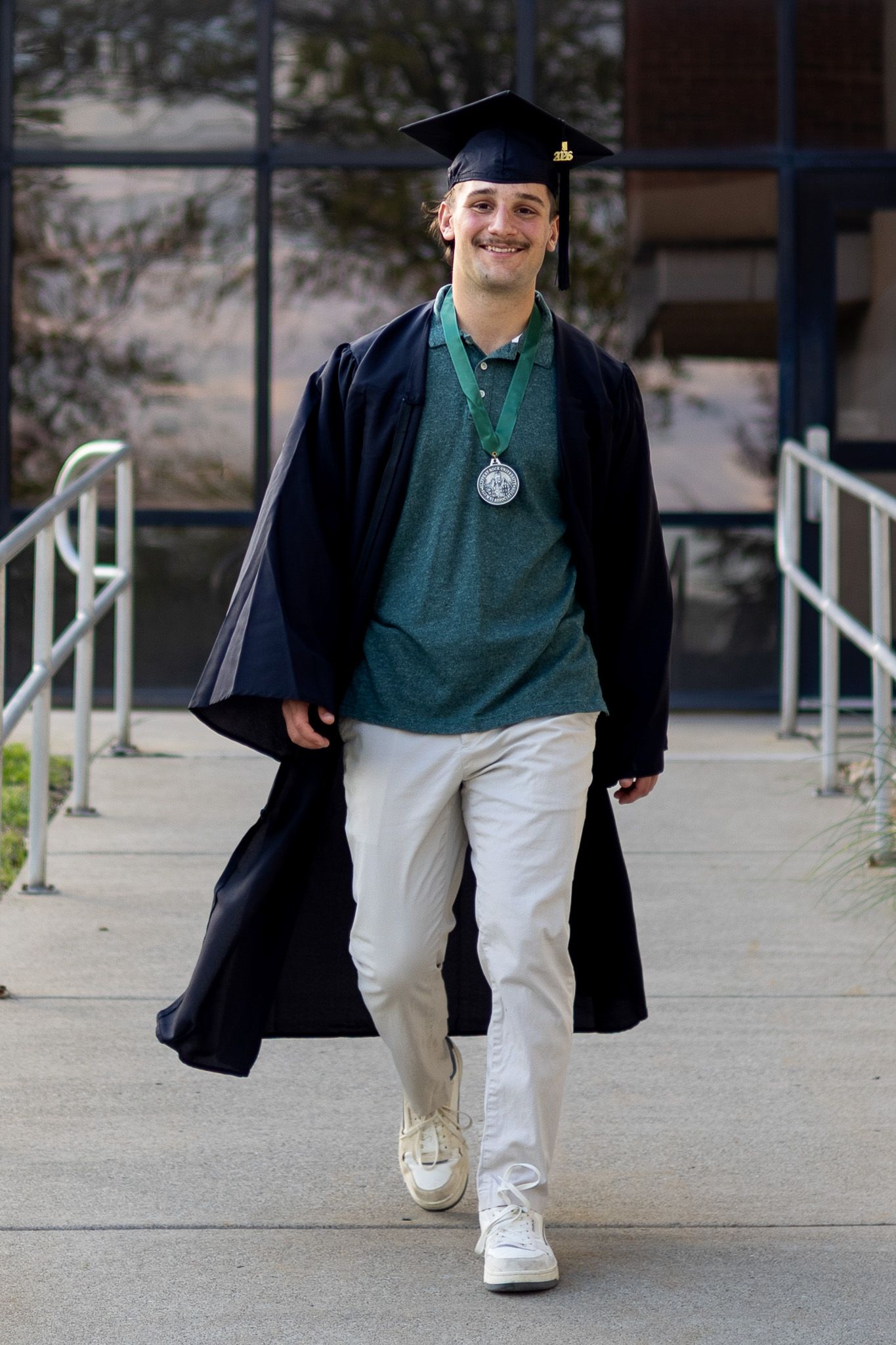 Male graduate walks in cap and gown with medal at Slippery Rock University campus.
